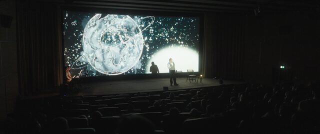 Blick vom Rang auf die Leinwand: ein leuchtender Planet, davor der Erzähler auf der Bühne
